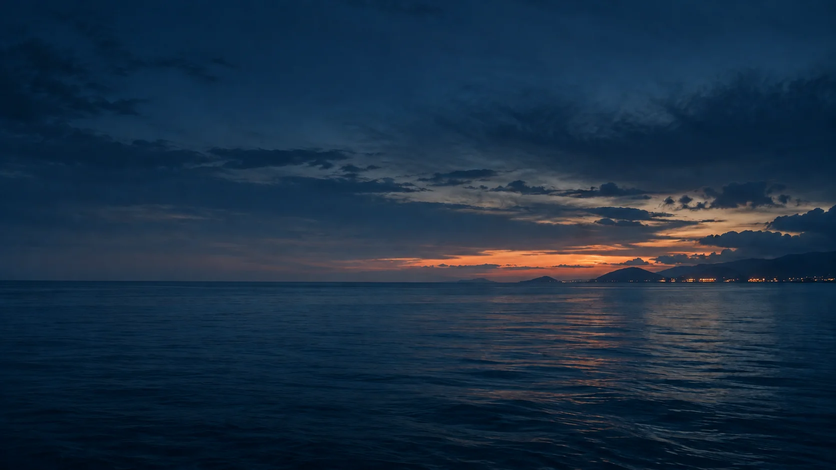 Calm open sea at dusk beneath blue clouds, with a distant coastline